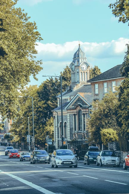 A street scene in Barnet featuring a large historic church with a prominent clock tower and a pointed spire, surrounded by trees with green foliage. Parked and moving cars line the road in front of the church, with some vehicles waiting at a traffic signal. The street has traffic lights, road markings, and pavement, and appears to be part of a typical residential and commercial area. In the foreground, blue and silver cars are visible, with a few pedestrians possibly nearby out of view. The sky is partly cloudy, with patches of blue sky and white clouds. This scene reflects a typical day in a UK town setting, aligned with house removals and moving logistics by Barnet Movers, where the surrounding environment and vehicle activity indicate urban planning and transportation processes involved in home relocation or furniture transport services.