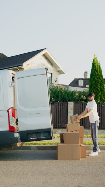 A young man in casual clothing is engaged in home relocation activities, standing outside a residential property next to a white moving van with its rear doors open. He is handling cardboard boxes, stacking and preparing them for loading into the vehicle. The boxes are of various sizes, made from brown corrugated cardboard, and some are sealed with packing tape. The scene takes place on a residential street, with a paved road, a dark wooden fence, and green shrubbery and trees, including a tall conifer, visible in the background. The house in the background has a pitched roof and white exterior walls. Inside the van, there are additional moving supplies, such as packing blankets, plastic wrap, and a trolley, which are partly visible through the open doors. This image illustrates the packing and loading process involved in furniture transport and home relocation, with [COMPANY_NAME] facilitating the move, consistent with the context of a house removals service as detailed in the Barnet removals cost guide.