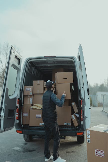 A man wearing a black cap, dark vest, and black trousers is inside the open rear of a white moving van, surrounded by numerous cardboard boxes of various sizes, some with visible labels, and packing m