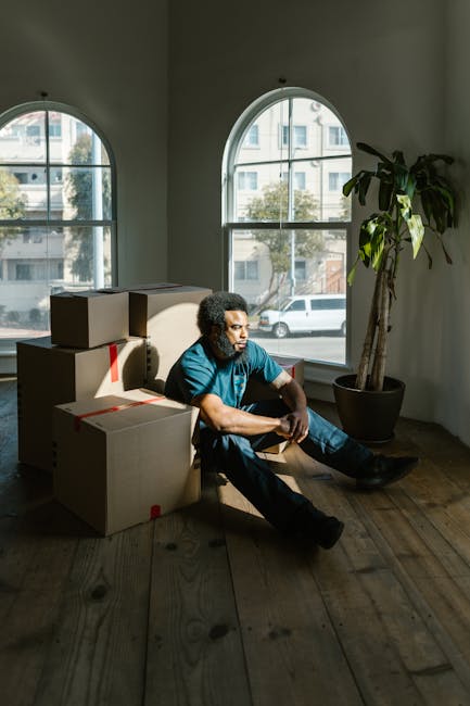 A man with curly dark hair and medium skin tone is sitting on the wooden floor inside an empty room with large, arched windows that have black frames, allowing natural daylight to illuminate the space. He is wearing a blue t-shirt and dark pants, resting against a stack of three cardboard boxes secured with red packing tape, which are placed near the corner of the room. The room features white walls, a potted plant with large green leaves situated to the right, and the windows reveal a view of a street with buildings and parked cars outside. The scene captures the moment during a home relocation or moving process, associated with the services of Barnet Movers, showing a pause amid furniture transport and packing activities involved in house removals or quick local moving services.