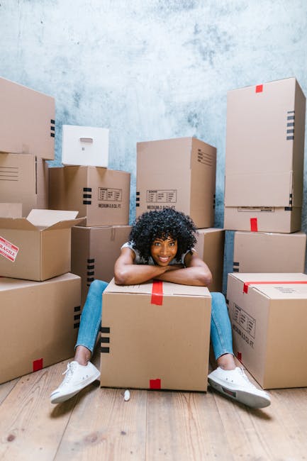 A young woman with curly black hair, smiling happily, is sitting on the wooden floor of a room surrounded by numerous cardboard boxes of varying sizes, some sealed with red or black tape. She is leaning forward with her arms resting on a large box in front of her, wearing a grey t-shirt, blue jeans, and white sneakers. The boxes are stacked along the walls and on the floor, with some partially opened, revealing packing materials inside. The room has a textured light blue wall and bright, natural lighting, suggesting an active home relocation or packing process. Barnet Movers' professional removal services are supported by the scene's depiction of packing and moving logistics, highlighting the preparation involved in furniture transport and house removals for a same-day local move in Barnet, EN5.
